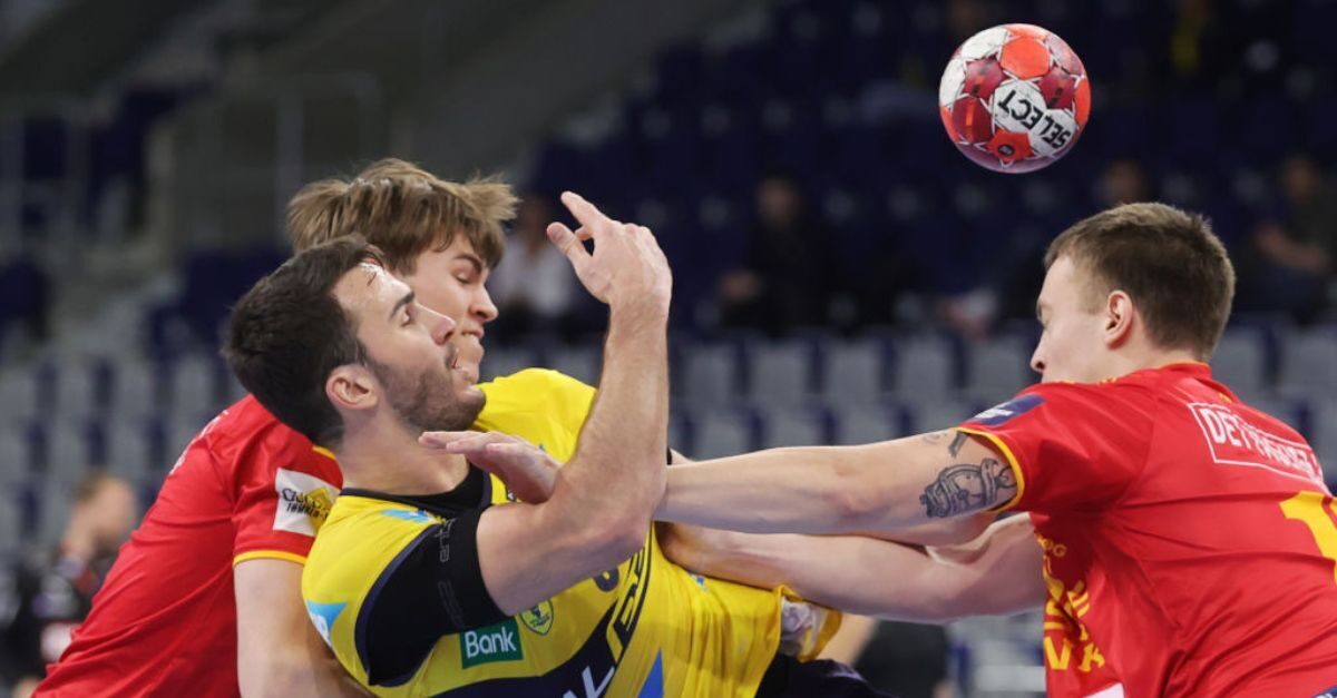 A handball player makes a move past a defender to try to score a goal while a teammate looks on.