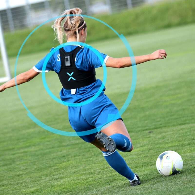 A women's soccer player is kicking the ball towards the goal while wearing a sports vest with a sensor that is compiling information about her performance.